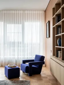 A modern living room with a blue upholstered armchair and footstool, white transparent curtains, and a wall-mounted shelf.