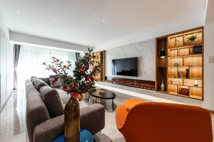 A modern living room featuring a grey sofa, orange chairs, pomegranate plant, glass vase, and wooden shelves with lighting.