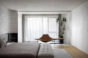 Minimalist bedroom with a wooden bed and a sleek, curved brown lounge chair in front of a large window with sheer curtains.