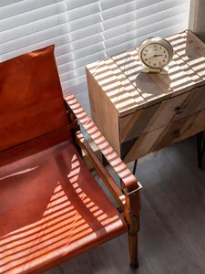 A stylish, industrial-style室内空间，配有棕色皮革扶手椅和木质抽屉柜，阳光透过百叶窗洒在椅面上。