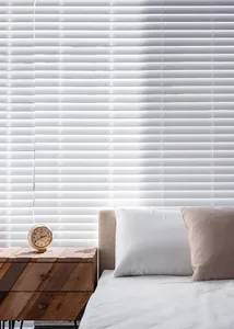 A minimalist bedroom featuring white blinds, a bed with white sheets and pillows, and a wooden side table with a clock.