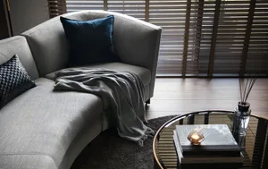 A modern living room with a grey couch, blue pillow, and wooden floor, featuring a glass coffee table and a window with blind