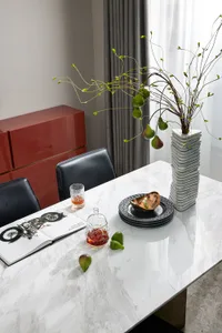 A modern dining room featuring a white marble table with black chairs, a vase with green figs, a glass of amber liquid, and a