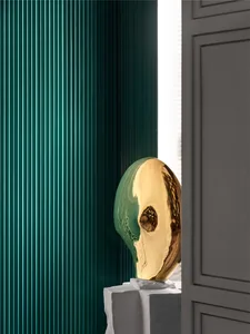 A modern, golden, sculptural object with a textured, reflective surface placed on a pedestal in a room with green vertical bl