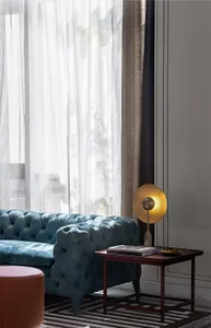 Modern living room featuring a blue upholstered sofa, sheer curtains, and a wooden side table with a lamp and books.
