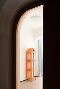 An orange metallic house-shaped shelf with three levels, standing in a corridor with a curved doorway and white curtains in t