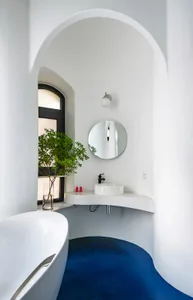 A minimalist bathroom with a white tub, blue flooring, a round mirror, and a potted plant.
