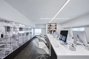A minimalist workspace featuring a row of white desks lined with white chairs and a clear glass partition