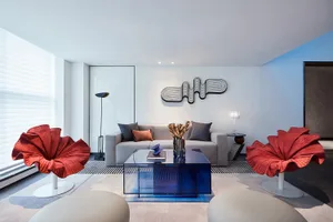 A modern living room features a sleek sofa with decorative pillows, minimalist red fabric chairs, a transparent blue coffee t