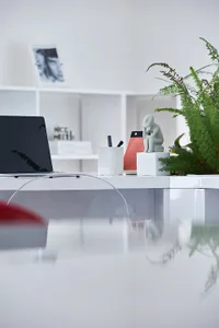An office workspace featuring modern white furniture, a clear glass desk, and a green plant.