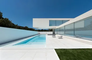 Modern minimalist residential exterior with concrete walls and large glass windows, featuring a sleek white patio area and a
