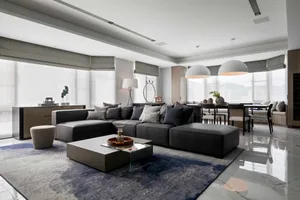 A modern living room with a gray sectional sofa, a low glass-top coffee table, and pendant lights. The room features large wi
