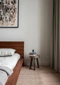 A minimalist bedroom featuring wooden furniture and a hanging light bulb on a stool, with a framed abstract painting above th