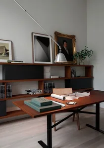 A modern home office desk with a wooden tabletop and metal legs, surrounded by shelves and framed art.