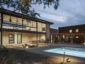 A modern two-story house with a brightly lit backyard featuring a pool, fire pit, and covered seating area.