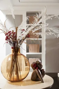 A round amber glass vase with dried white flowers and red berries sits on a circular table.