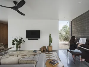 A living room with a minimalist design featuring concrete walls, a large gray sofa, a cactus plant, and a person playing the 