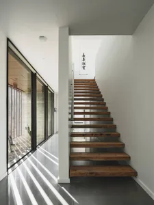 A minimalist house interior featuring a modern wood and glass staircase with sunlight streaming in through the large windows.