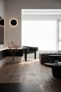 A modern minimalist living room featuring wooden floors, black furniture, and a large window with blinds.