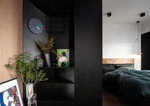 A modern bedroom featuring a dark-colored shelf with a clock, plants, and decor, leading to a bed with velvet bedding and a w