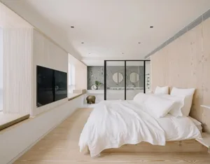 A minimalist bedroom with wooden flooring and a white bedspread, featuring a bathroom in the background with circular mirrors