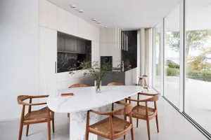 A modern residential kitchen featuring a white marble table and wooden chairs, with a view of the outdoors through large glas