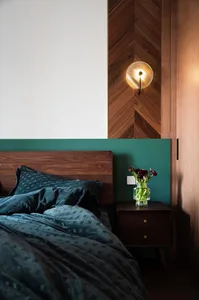 A modern bedroom with a wooden headboard and green accents, featuring a bedside table with a vase of flowers and a wall-mount