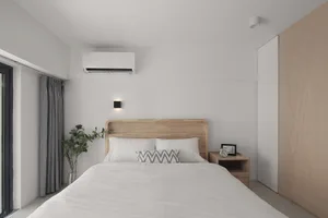 A minimalist bedroom features a wooden bed with a white headboard and footboard, surrounded by a simple, white bedspread and