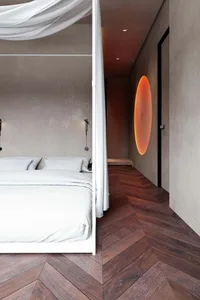 A modern minimalist bedroom featuring a white bed canopy, herringbone wood floor, and a round orange light fixture on the bei