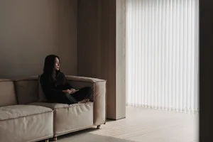 A person sitting on a modern living room leather couch looking out of a window with vertical blinds.