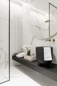 A minimalist bathroom with white marble walls and floor, black floating vanity, and round mirror.