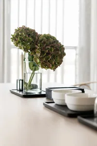 A minimalistic table setting with a clear glass vase containing hydrangea flowers and bowls on a tray.