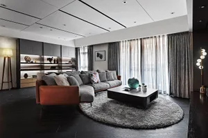 A modern living room with dark flooring, a large grey sofa with patterned cushions, a minimalist coffee table, and grey curta