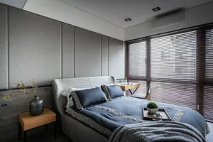 A modern minimalist bedroom featuring grey upholstered walls, wooden furniture, and a small wooden desk with a plant and cup.