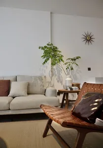 A minimalist living room with a beige sofa adorned with cushions, a wooden leather chair, and a small wooden side table with 