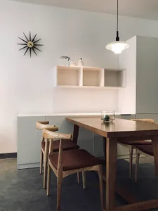 Modern minimalist dining room with wooden table and chairs against a white wall, featuring a unique sunburst clock and minima