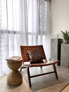 A modern living room featuring a wood and leather chair, white sheer curtains, and a concrete pedestal with a plant and fishb