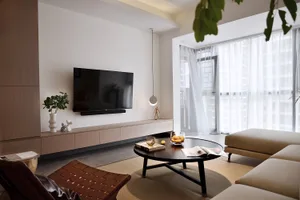 A modern living room featuring a flat-screen TV, round wooden coffee table, beige sofas, and a brown woven chair with a white