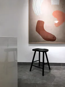 A modern art gallery exhibit featuring a black stool on a concrete floor, with an abstract painting hanging on the wall.