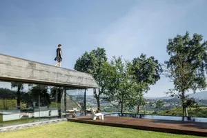 A modern house with a concrete roof and glass walls, featuring a person walking on the roof against a clear blue sky.