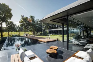 A modern house with a flat roof, glass walls, and concrete patio featuring a swimming pool and outdoor seating area.