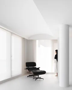 A minimalist white interior with a sleek, modern chair and a person standing in a white room with large windows.