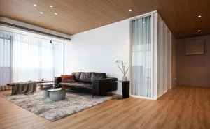 A contemporary living room with a black leather sofa, wooden flooring, and large floor-to-ceiling windows.