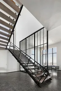 A modern metal staircase with a glass wall and wooden steps, set against a white wall and black floor.