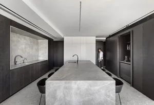 A modern residential kitchen with a long marble island centerpiece, dark wooden cabinetry, and sleek black chairs.