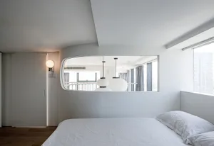Modern minimalist bedroom with white walls, bed, and furniture, featuring a unique curved mirror and white lighting fixtures.