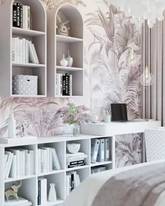 A contemporary home office features blush palm wallpaper, modern furniture, and white bookshelves filled with books and decor