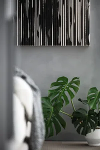 A monstera plant in a concrete planter with a canvas artwork featuring vertical black and white lines hanging on a matte gray