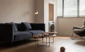 A modern living room featuring a wooden floor, a dark gray sectional sofa, a coffee table, a standing lamp, and a window with