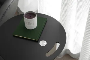 A modern side table with a white ceramic candle holder, a green hardcover book, and a white textured curtain in the backgroun